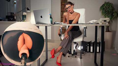Media: Video of a professional woman in glasses, wearing a black top and grey pants, sitting at a desk in an office, with a dildo in a glass ball.