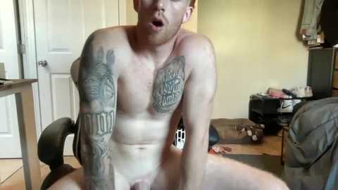 Video of a shirtless, muscular, fair-skinned man with a beard and tattoos on his chest and arms, sitting on a floor in a cluttered room.