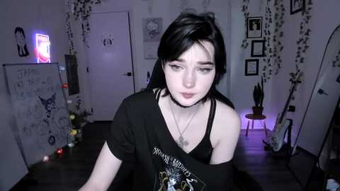 A video of a young, pale-skinned woman with black hair, wearing a black off-shoulder shirt and choker, standing in a dimly lit, gothic-themed bedroom with dark walls, framed art, and neon lights.