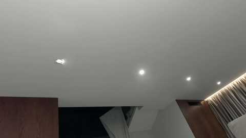 A video of a modern, minimalist room with a white ceiling featuring three recessed lights, a wooden wall, and a sleek, white curtain rod.