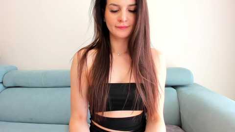 Media: Video of a young woman with long, straight brown hair, wearing a black strapless crop top, seated on a light blue sofa in a minimalist room.