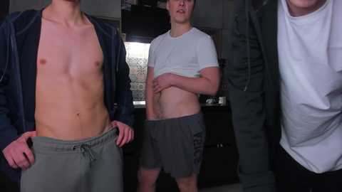 Video of three shirtless young men in casual attire; one in a dark jacket, another in a white shirt, and the third in gray shorts, standing in a dimly lit kitchen.