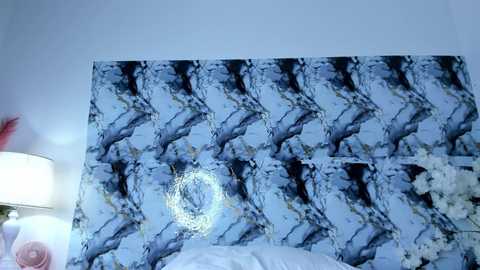 Video of a bedroom wall featuring a marble-patterned wallpaper with a circular, metallic, textured centerpiece. The scene includes a partially visible lamp and a flower arrangement in the background.