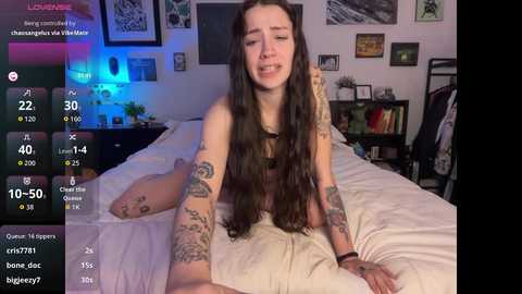 Media: Video of a young, pale-skinned woman with long dark hair, smiling while lying on a white bed, surrounded by colorful wall art and a green plant, in a cozy bedroom.