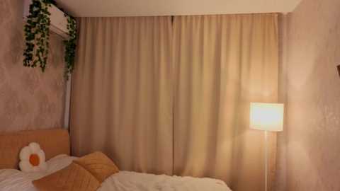 Video of a cozy bedroom with beige curtains, a white floor lamp, and a bed with white sheets and brown pillows.