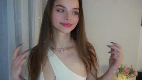 Video of a young woman with long brown hair, fair skin, and a slender physique. She wears a white dress with a single strap and a silver necklace, smiling slightly. Background includes a light-colored wall and a floral arrangement.