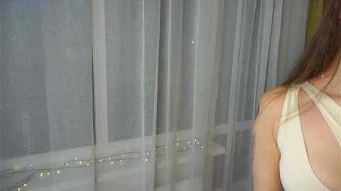 A video of a woman with long brown hair, wearing a sleeveless white top, standing in front of sheer white curtains adorned with string lights.