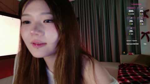 Media: Video of an Asian woman with long brown hair, fair skin, and a neutral expression, wearing a white top, in a dimly lit room with a digital overlay of social media metrics.