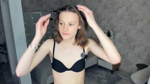 Video of a slender, Caucasian woman with shoulder-length brown hair, fair skin, wearing a black bra and silver jewelry, adjusting her hair in a dimly lit, gray-toned room with concrete walls.