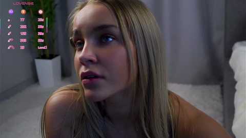 Media: A video of a blonde woman with fair skin and blue eyes, looking contemplative, in a dimly lit room with a plant and a bed in the background.