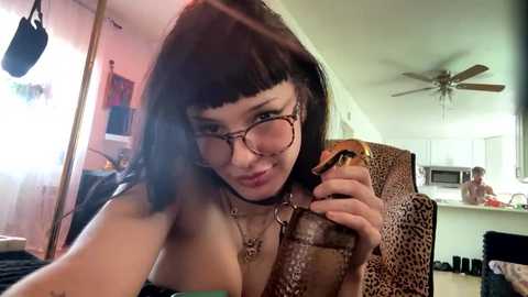 Video of a young woman with fair skin, straight black hair, and glasses, wearing a leopard-print top, holding a hot dog in a dimly lit living room.