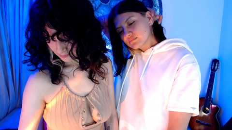 Video of two women with curly dark hair, one in a beige dress, the other in a white hoodie, standing close together, against a blue-lit background, with a guitar on the right.