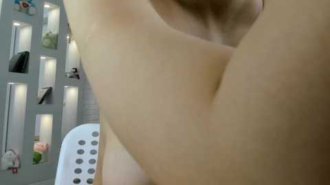 Video of a nude person with light skin, lying on their back on a white plastic chair, in a modern, well-lit room with white shelving units and small decorative objects.