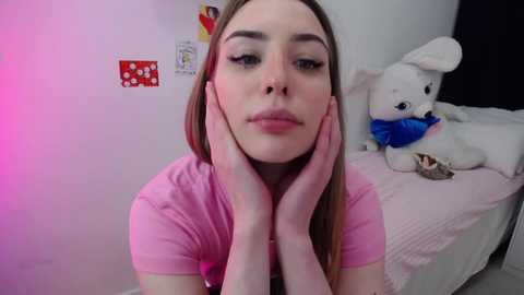 Media: Video of a young woman with fair skin, straight blonde hair, and blue eyes, wearing a pink t-shirt, resting her hands on her face, in a pink room with a plush rabbit on a bed.