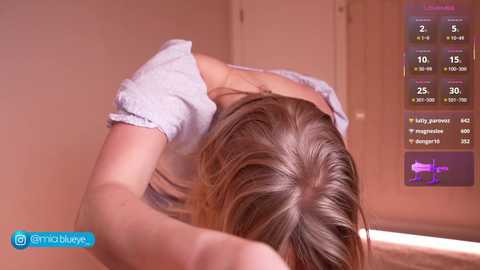 Video of a blonde woman with fair skin, wearing a white t-shirt, leaning over a table with her head resting on her arms, in a dimly lit room.