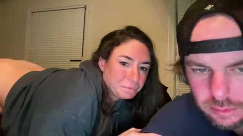 Video of a young woman with long dark hair, wearing a black jacket, and a man in a black cap, both on all fours, in a dimly lit room.