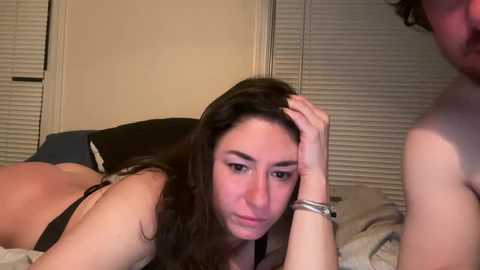 Video of a young woman with long brown hair, lying on a bed, wearing a black bra, looking tired and holding her head.