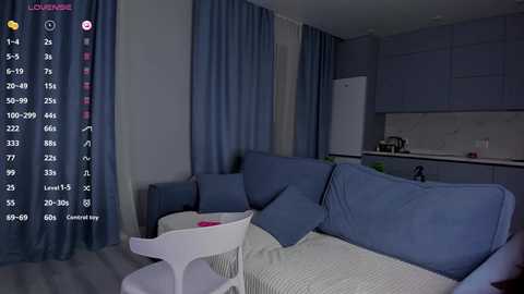 Video of a modern, minimalist living room with light blue sofas, white chairs, and grey cabinets. A fridge and kitchenette are visible in the background.