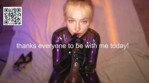 Media: Video of a blonde woman in a shiny black latex bodysuit, kneeling with a black dildo in her mouth, on a bed with white sheets. QR code in the top left corner.
