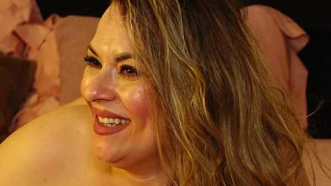 Video of a smiling, middle-aged woman with light skin and wavy, shoulder-length brown hair. She wears red lipstick and has a radiant complexion. The background is dimly lit, featuring soft, pink pillows.