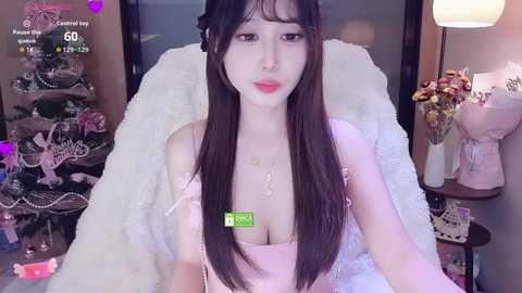 Media: A video of a young Asian woman with long, straight black hair and fair skin, wearing a pink off-shoulder top, sitting on a white fur rug in a cozy, dimly lit room.