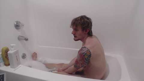 Video of a fair-skinned man with short, messy brown hair, sitting shirtless in a white bathtub filled with bubbles, showcasing a colorful tattoo on his left arm, surrounded by white walls and bathroom items.