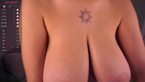 Video of a nude woman with large, natural breasts featuring a small sun tattoo on her upper chest. Background includes a black couch and a digital clock display.