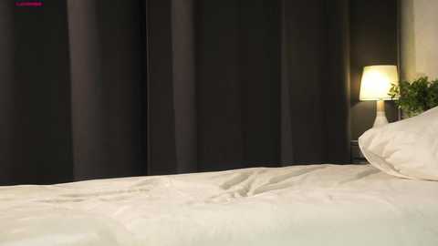 Video of a neatly made bed with white linens, a dark curtain backdrop, and a small table lamp emitting a warm light.