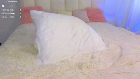 A video of a plush, white, tufted bed with a fluffy, cream-colored blanket, adorned with a large, white pillow. The headboard is beige and pink throw pillows are placed on either side.