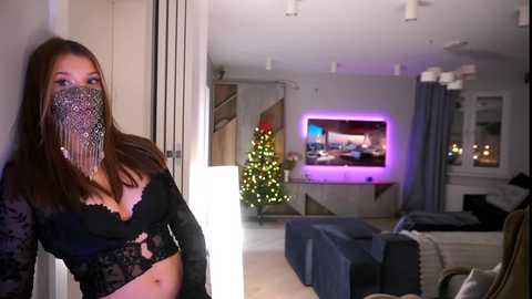Media: A video of a young woman with long brown hair, wearing a black lace bra, standing in a modern living room with a Christmas tree and TV.