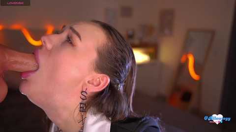 Video of a pale-skinned woman with dark hair, wearing a white blouse and black earrings, performing oral sex on a man with a visible erection. Background is blurred, showing a dimly lit room with a TV and some decor.