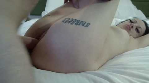Video of a naked woman with pale skin, small breasts, and a tattoo reading \"RAPPER\" on her hip, lying on a white bed.
