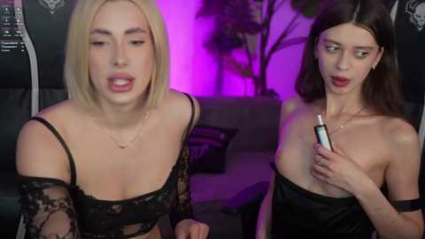 Video: Two women, one blonde in black lace, the other brunette in a strapless black dress, sitting, holding a lighter, purple lighting, indoor setting.