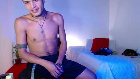 Media: Video of a shirtless, fit, tattooed young man with medium skin tone, wearing black shorts, smiling, in a dimly lit bedroom with white walls, red pillows, and a blue light.