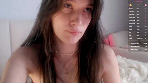 A video of a young woman with long, dark hair, fair skin, and a delicate facial structure, wearing a beige top and necklace. She has a neutral expression and sits on a white couch in a softly lit room.