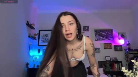 Video of a tattooed woman with long dark hair, wearing a white bra and black pants, leaning forward in a dimly lit room with purple and blue LED lighting, framed art on the walls, and a bed in the background.
