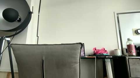 Video of a minimalist room with a large, black, round studio light on the left, a gray cushioned chair in the center, a pink cloth on a black desk to the right, and a mirror reflecting a bottle.