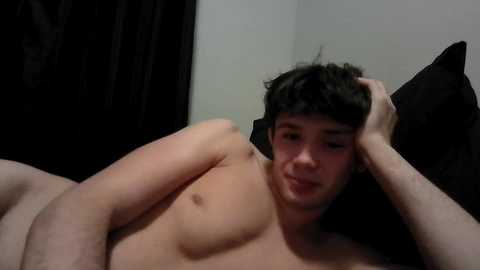 Video of a shirtless, young, Caucasian male with dark hair, lying on a bed with black sheets, looking tired and holding his head.