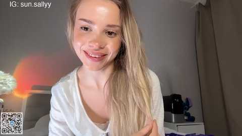 Video of a smiling, fair-skinned, blonde young woman with long hair, wearing a white blouse, in a softly lit bedroom.