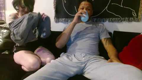 Video of a man in gray pajamas drinking from a blue cup, while a woman in a gray shirt and black shorts kneels beside him, both sitting on a black couch.