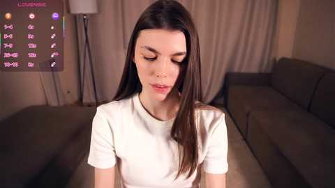 Media: Video of a young Caucasian woman with long brown hair, wearing a white t-shirt, in a dimly-lit living room with a sofa, a lamp, and a digital clock displaying the time.