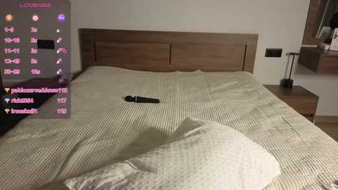 A video of a modern bedroom with a beige, textured bedspread, wooden headboard, and a small black vibrator on the bed. The room features a minimalist design with a nightstand on the right.