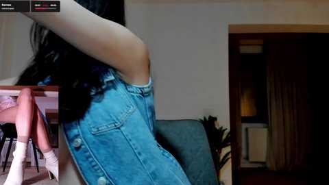 Video of a person wearing a denim overalls, captured from a low angle, with a view of a dimly lit room featuring a sofa and a partially visible window.