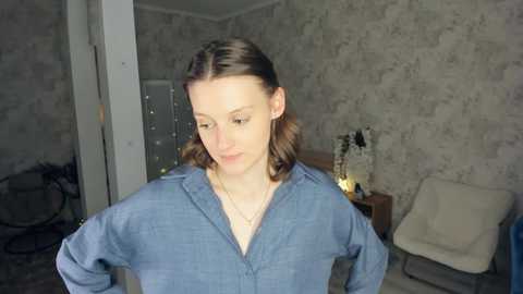 Media: Video of a young woman with shoulder-length brown hair, wearing a blue button-up shirt, standing indoors in a dimly lit room with a textured gray wall, a white chair, and a wooden nightstand with a lit candle.