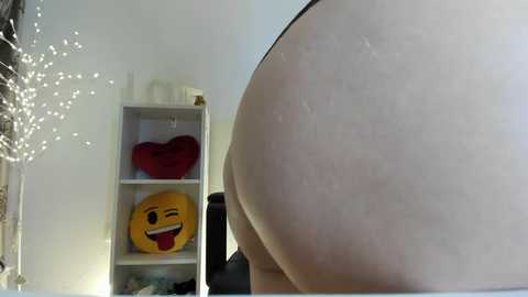 A video showing a close-up of a large, smooth buttock, likely female, in a bedroom. Background features a white shelf with a red heart pillow and a yellow smiley face pillow.