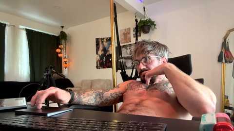 Video of a shirtless, tattooed, muscular man with glasses, wearing a black wristband, using a laptop in a cluttered, brightly-lit room with posters and a green plant.