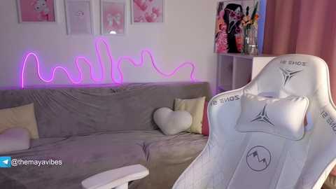 A video of a cozy living room with a gray sectional sofa adorned with heart pillows and a neon \"Mama\" sign. A white gaming chair with \"SMX\" branding is prominently featured in the foreground.