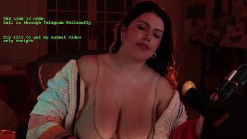 Video of a curvy, light-skinned woman with long, dark hair and large breasts, wearing a low-cut top and knit cardigan, sitting at a desk with a microphone, in a dimly lit room with posters and shelves in the background.