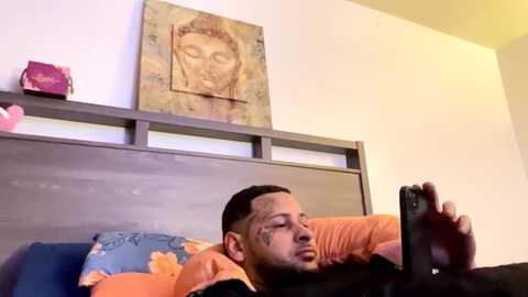 Media: Video of a young Black man with a beard, wearing an orange shirt, lying on a bed with a blue pillow, holding a smartphone, in a modern bedroom with a wooden headboard and Buddha painting.