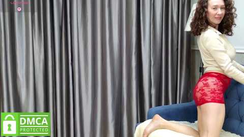 Media: Video of a smiling, curly-haired woman in a beige sweater and red lace shorts, kneeling on a blue chair in front of grey curtains.